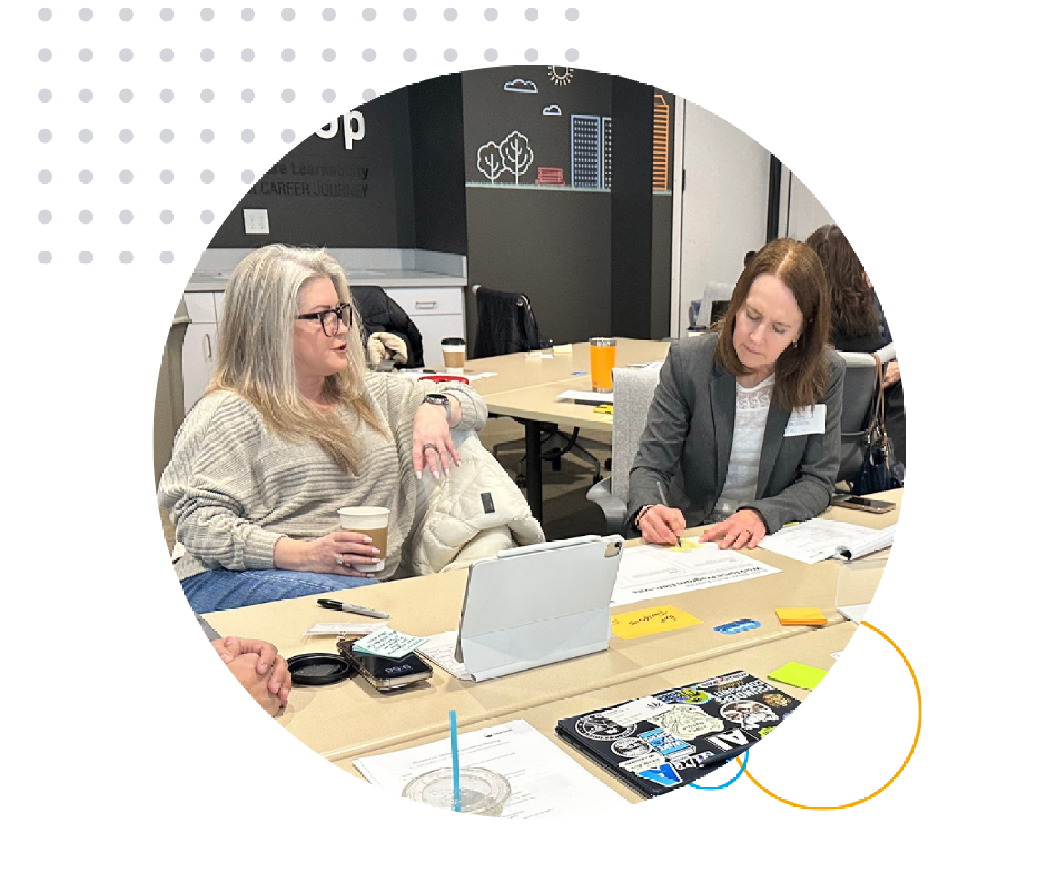 Two people are seated at a table in an office, discussing notes. One holds a coffee cup, the other writes on paper. A laptop and post-its are on the table.