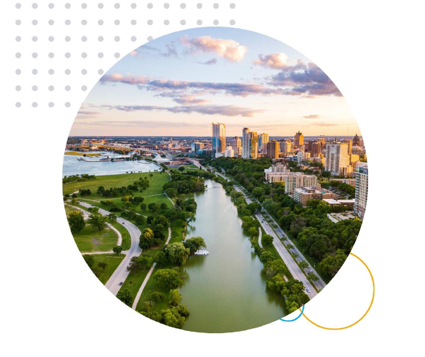 Aerial view of Milwaukee cityscape at sunset with a river surrounded by green parks, trees, and modern high-rise buildings, set against a dramatic sky.